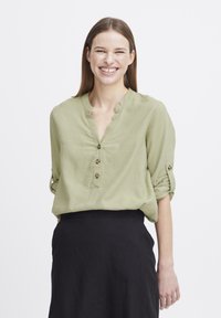 Blouse verte claire à col, avec des manches retroussées et trois boutons de fermeture ; présente une texture douce et une coupe décontractée, associée à une jupe noire.