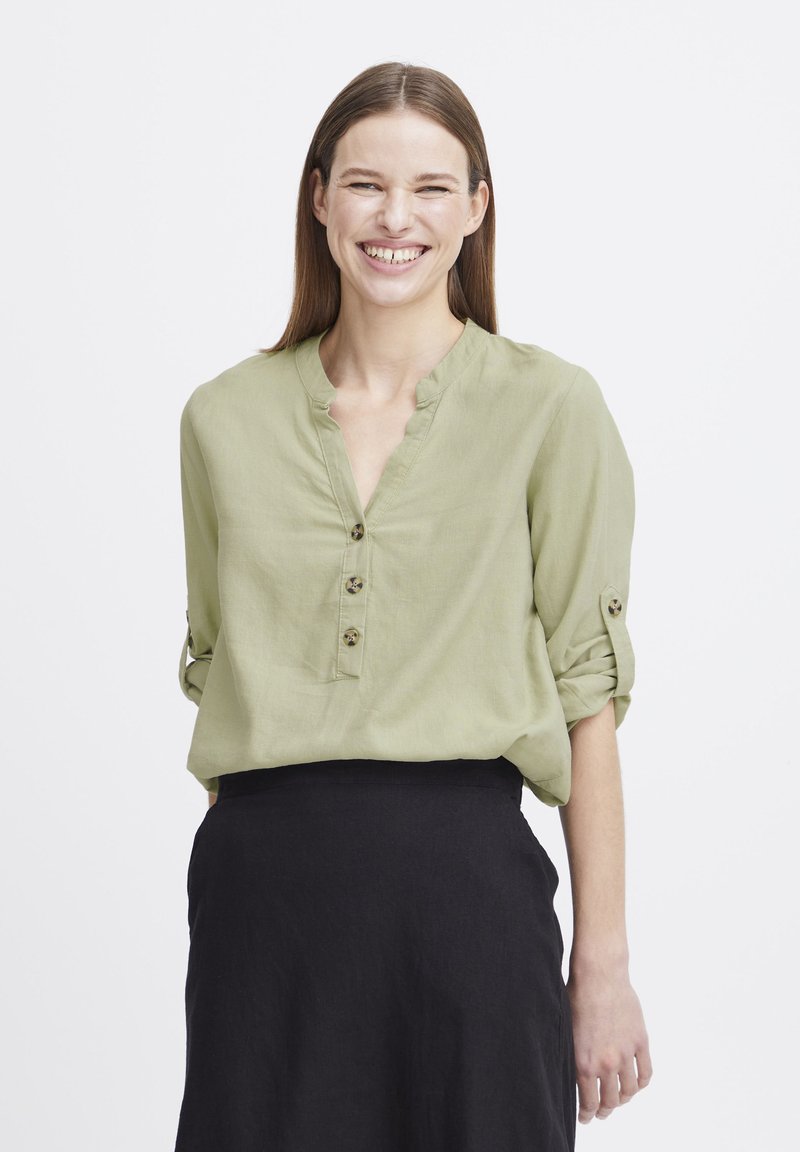 Blouse verte claire à col, avec des manches retroussées et trois boutons de fermeture ; présente une texture douce et une coupe décontractée, associée à une jupe noire.