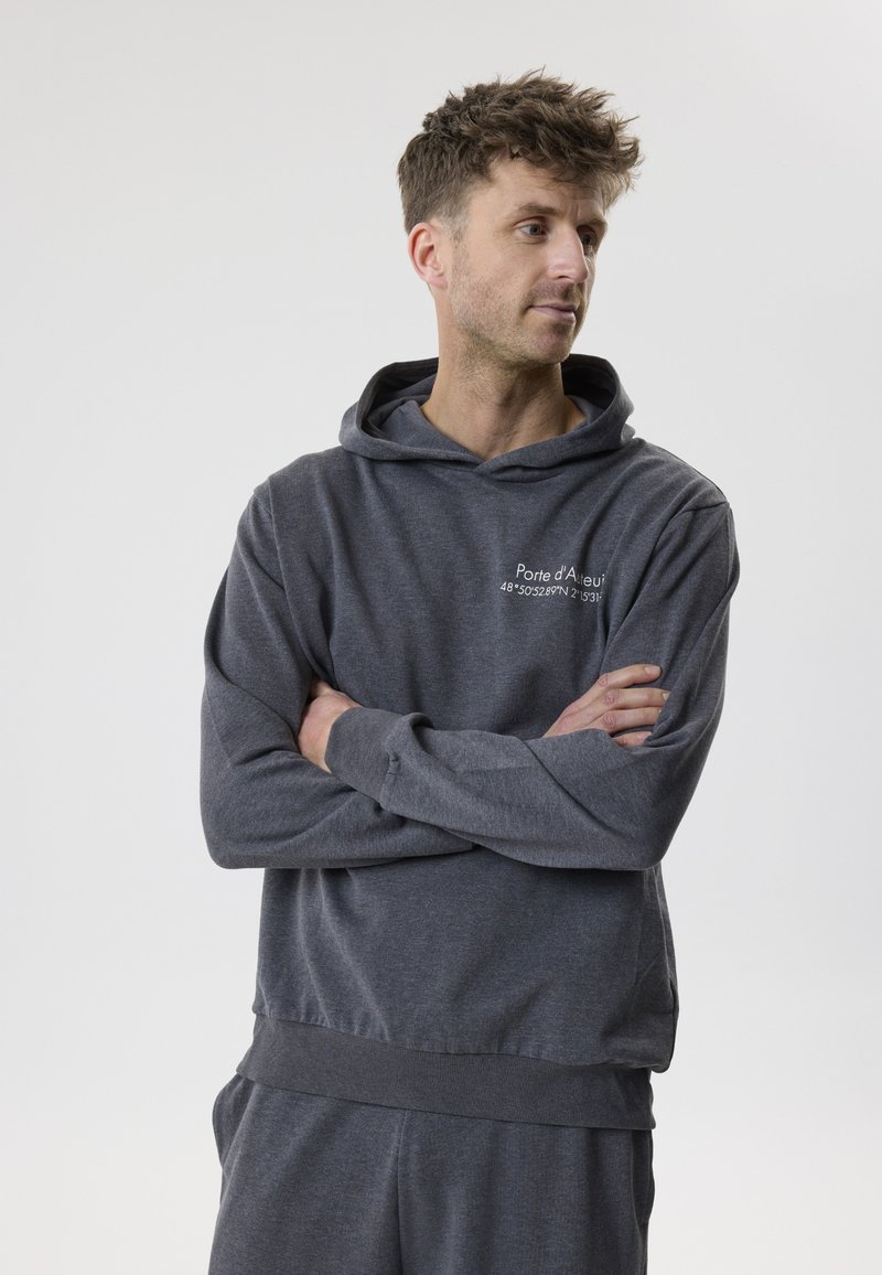 Homme aux cheveux courts portant un sweat à capuche gris foncé et un pantalon assorti, bras croisés, regardant vers la droite sur un fond clair uni.