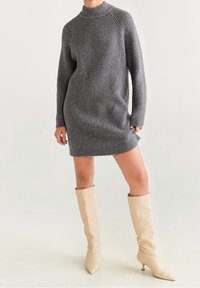 Šedé pletené šaty se stojáčkem a dlouhými rukávy, doplněné krémovými kozačkami po kolena s špičatou špičkou.