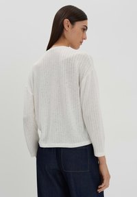 Loose-fitting white knit sweater with a ribbed texture, long sleeves, and a rounded hem, paired with dark denim trousers.