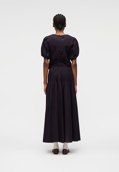 Femme aux cheveux tressés vue de dos, portant un haut noir à manches bouffantes et une longue jupe, avec des chaussures marron et des chaussettes blanches.