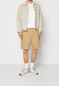Giacca beige sopra una t-shirt bianca, abbinata a pantaloni corti khaki. Sneakers grigie con accenti rosa, calze bianche. Tessuto morbido, design casual.