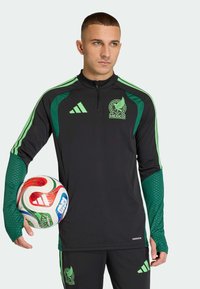 Fekete, zöld díszítésekkel és Mexikó címerével ellátott sportos cipzáras ing, hozzáillő nadrággal. A játékos színes focilabdát tart a kezében.