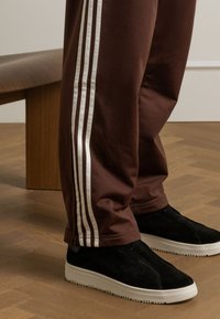 Braune Jogginghose mit weißen Seitstreifen, locker geschnitten und mit Reißverschluss an den Knöcheln. Kombiniert mit schwarzen Wildleder-Slippern mit weißen Sohlen.