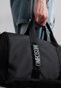 Ręka trzyma czarną torbę sportową Moschino, widoczna osoba ma na sobie jasnoniebieskie dżinsy i kurtkę, widok od pasa w dół.