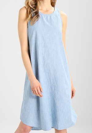 Femme portant une robe sans manches, bleu clair, longueur genou, avec de subtiles rayures verticales, sur un fond uni.