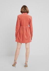 Šaty z kordu v rezavé barvě s dlouhými rukávy, nařaseným pasem a mírně rozšířenou sukní. Nosené s sandály na vysokém podpatku a tenkými pásky.