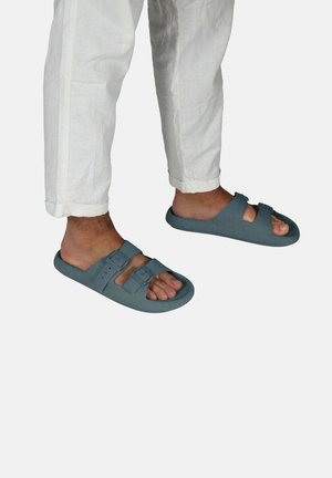 Claquettes en caoutchouc bleu avec deux sangles ajustables et boucles, présentant un design à bout ouvert et une texture lisse, portées avec un pantalon blanc.