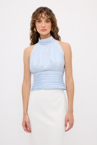 Top a halter blu chiaro con pieghe verticali e texture arricciata, vita aderente e collo alto. Tessuto liscio, abbinato a una gonna bianca.