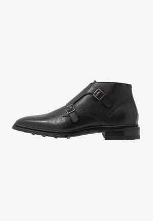 Bottine en cuir noir avec boucles double monk strap, bout arrondi et talon empilé sur fond blanc.