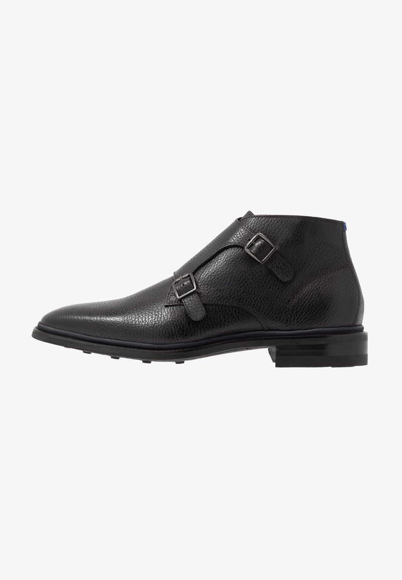Bottine en cuir noir avec boucles double monk strap, bout arrondi et talon empilé sur fond blanc.