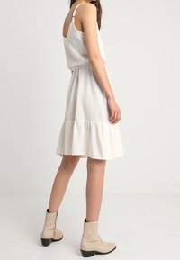 Vit, ärmlös klänning med en lager-på-lager nederkant, smal midja och mjukt tyg. Bärs med beige ankla stövlar med svarta klackar.