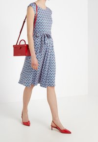 Niebieska sukienka w wzory z krótkimi rękawami i tied w talii, zestawiona z czerwoną torebką i dopasowanymi butami na obcasie typu slingback.