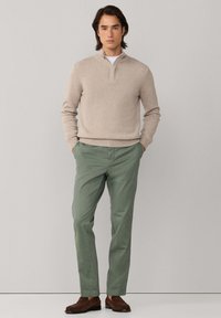 Jonge man staand met handen in zakken, gekleed in een beige trui met kwart-ritssluiting, wit ondershirt, olijfgroene broek en bruine loafers, tegen een effen muur.