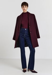 Cappotto di lana bordeaux, maglione a maglia bordeaux, jeans a zampa d'elefante in denim scuro e tacchi a punta scura. Presenta bottoni e cuciture a contrasto.