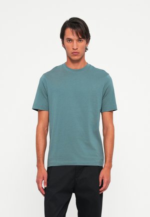 Man wearing a teal short-sleeve t-shirt and black pants, standing against a plain white background, facing forward.