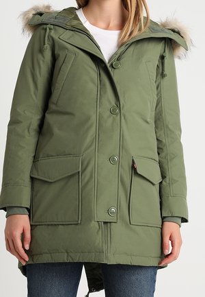 Frau trägt eine olivgrüne Winterparka mittlerer Länge mit Knopf- und Reißverschluss, zwei großen Fronttaschen mit Klappen und einer mit Pelz besetzten Kapuze.