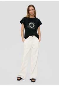 Schwarzes Baumwoll-T-Shirt mit kurzen Ärmeln und weißem Text, kombiniert mit hellbeigen, hochgeschnittenen, weit geschnittenen Hosen und schwarzen Plateausandalen.