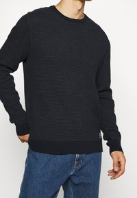 Schwarzer, strukturierter Pullover mit Rippmuster, Rundhalsausschnitt und langen Ärmeln. Mit einem einfarbigen schwarzen Saum und Bündchen.