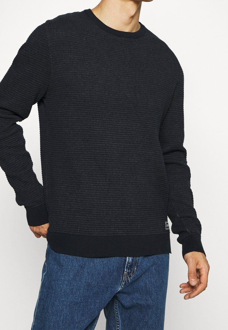 Schwarzer, strukturierter Pullover mit Rippmuster, Rundhalsausschnitt und langen Ärmeln. Mit einem einfarbigen schwarzen Saum und Bündchen.