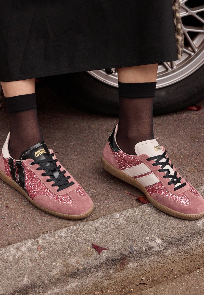 Pieds portant des baskets roses à paillettes avec des lacets noirs et des chaussettes noires aux chevilles, debout sur le trottoir près d'un pneu de voiture.