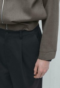 Braune Jacke mit Struktur und Reißverschluss, kombiniert mit dunkler Hose mit Knopfverschluss und plissiertem Design. Nahaufnahme einer Hand auf der Hose.