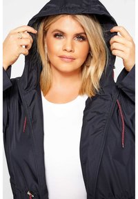 Kevyt, tummansininen vedenpitävä takki, jossa on huppu, säädettävät nyörit ja punaiset yksityiskohdat, pukeutuneena valkoisen paidan päälle.