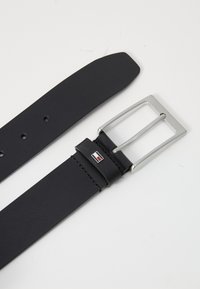 Cinturón de cuero negro con una hebilla rectangular de metal plateado, que presenta detalles de costura y un acento de marca cerca de la hebilla.