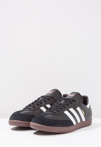 Un paio di sneakers Adidas Samba nere con strisce bianche, lacci neri, suole gomma e il testo dorato "SAMBA" sul lato, mostrati su una superficie bianca.