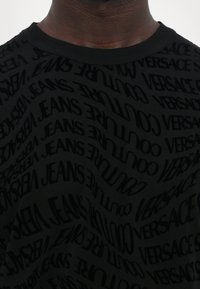 Juodas marškinėlis su pakartojančiu „Versace Jeans“ tekstu įvairiomis orientacijomis. Gumačiau apykaklė ir minkšta tekstūra.