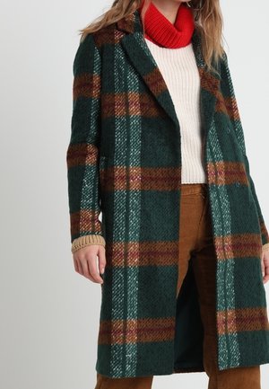 Kvinde iført en lang grøn- og brunternet frakke over en hvid sweater med en rød rullekrave og brune bukser.