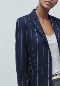 Granatowa marynarka w paski z dopasowanym krojem, z pojedynczym zapięciem na guzik, klapami z wycięciem i kieszenią na piersi. Teksturowana tkanina.