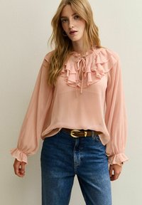 Blusa color durazno con cuello rizado, mangas largas y puños elásticos; combinada con jeans azul de tiro alto y un cinturón marrón.