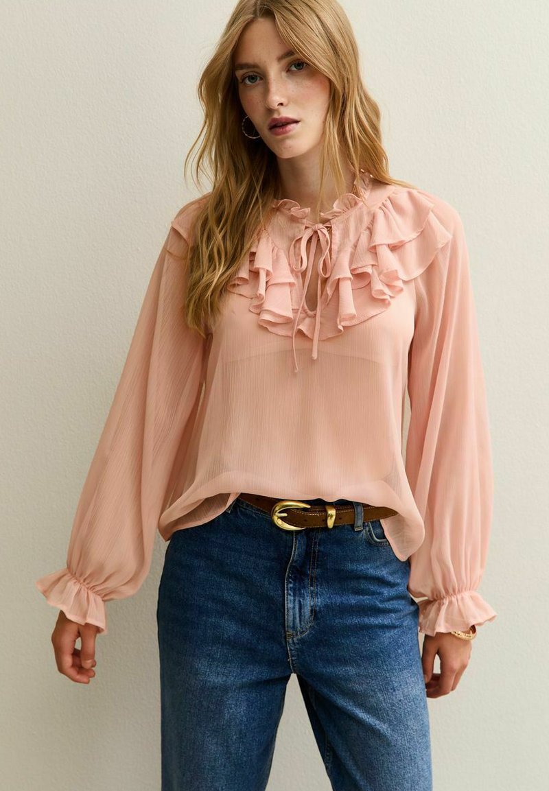 Blusa color durazno con cuello rizado, mangas largas y puños elásticos; combinada con jeans azul de tiro alto y un cinturón marrón.