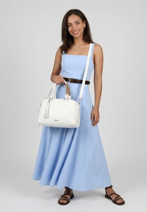 Donna con un vestito azzurro senza maniche e cintura marrone, che tiene una borsa bianca e indossa sandali marroni, sorridente su uno sfondo uniforme.