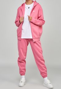 Roze hoodie en bijpassende joggers van zacht materiaal. Een wit T-shirt eronder met een klein zwart logo. Witte sneakers met enkelsokken.