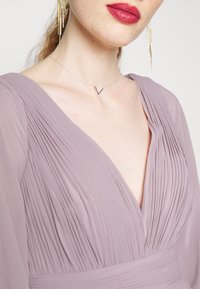 Robe plissée lavande avec un décolleté en V profond et des manches fluides, associée à un collier chaîne argentée avec un pendentif en forme de V.