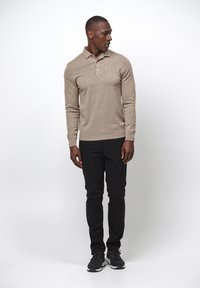 Beige Langarm-Polo mit einer Zwei-Knopf-Leiste, kombiniert mit schwarzen Slim-Fit-Hosen und dunklen Sneakers vor einem schlichten Hintergrund.