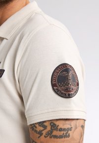 Jasbeżowa koszulka polo z teksturowanym materiałem, z okrągłą, brązową, wyszywaną naszywką na lewym rękawie, zawierającą szczegóły emblematu.
