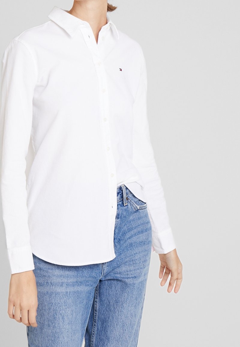 Personne portant une chemise blanche à manches longues boutonnée rentrée dans un jean en denim bleu sur un fond clair uni.