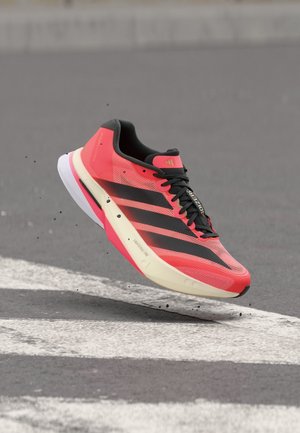 Scarpa da corsa corallo brillante con strisce e lacci neri, sospesa sopra una strada asfaltata con linee bianche dipinte e piccole particelle di polvere.