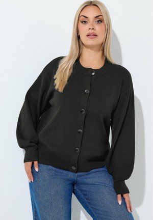Kobieta z długimi blond włosami, ubrana w czarny kardigan z guzikami i niebieskie jeansy, stojąca na jednolitym jasnym tle.