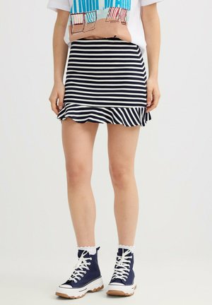 Mujer que lleva una minifalda con rayas horizontales en azul marino y blanco con dobladillo con volantes, zapatillas deportivas altas en azul marino y una camiseta corta con temática de playa.