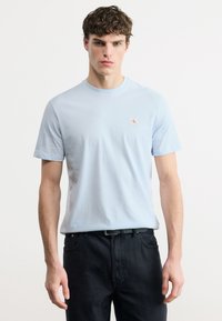 Νέος άνδρας που φοράει ένα ανοιχτό μπλε t-shirt Calvin Klein και μαύρο παντελόνι, στέκεται μπροστά σε ένα απλό λευκό φόντο.
