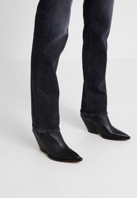 Botas de tobillo de cuña de cuero negro con punta afilada y superficie texturizada, combinadas con unos jeans de denim oscuros.