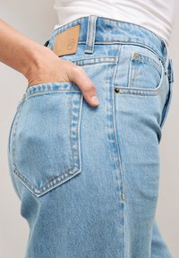 Jean en denim bleu clair avec taille haute, couture visible et patch en cuir à l'arrière. Présente une poche avec une main insérée.