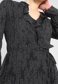 Vestido negro con líneas verticales discontinuas blancas, escote en V con volantes, mangas largas y una cinta en la cintura, llevado por una persona que se toca el cuello.