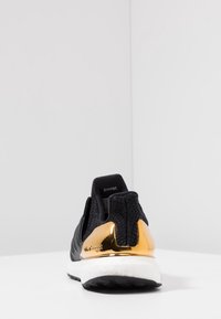 Černá sportovní obuv s horní částí z síťoviny, zlatě akcentovaný opatek, bílou mezipodešví boost a texturovanou gumovou podrážkou. Pouze zadní pohled.