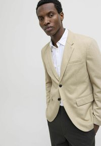 Beige blazer with a textured fabric, notched lapels, two front pockets, and black buttons, layered over a white shirt.